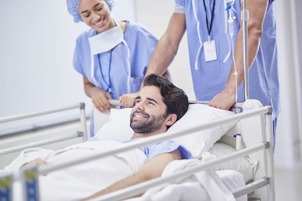 A smiling male patient lying on a hospital gurney being moved by two smiling healthcare workers in blue scrubs. The gurney has an IV pole attached with a visible IV bag and tubing.