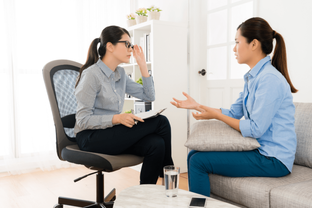A photo showing a counseling or therapy session between two women in an office setting, with one woman in an office chair holding a clipboard and the other woman seated on a couch gesturing as she speaks.