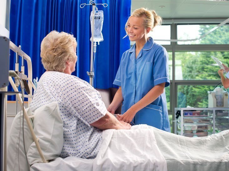 A young, smiling nurse in blue scrubs holding the hands of an elderly patient sitting in a hospital bed with an IV drip.