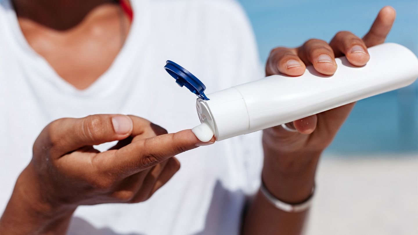A close-up photo of a person's hands, showing sunscreen being squeezed from a white bottle onto their fingertip, illustrating the application of sun protection.