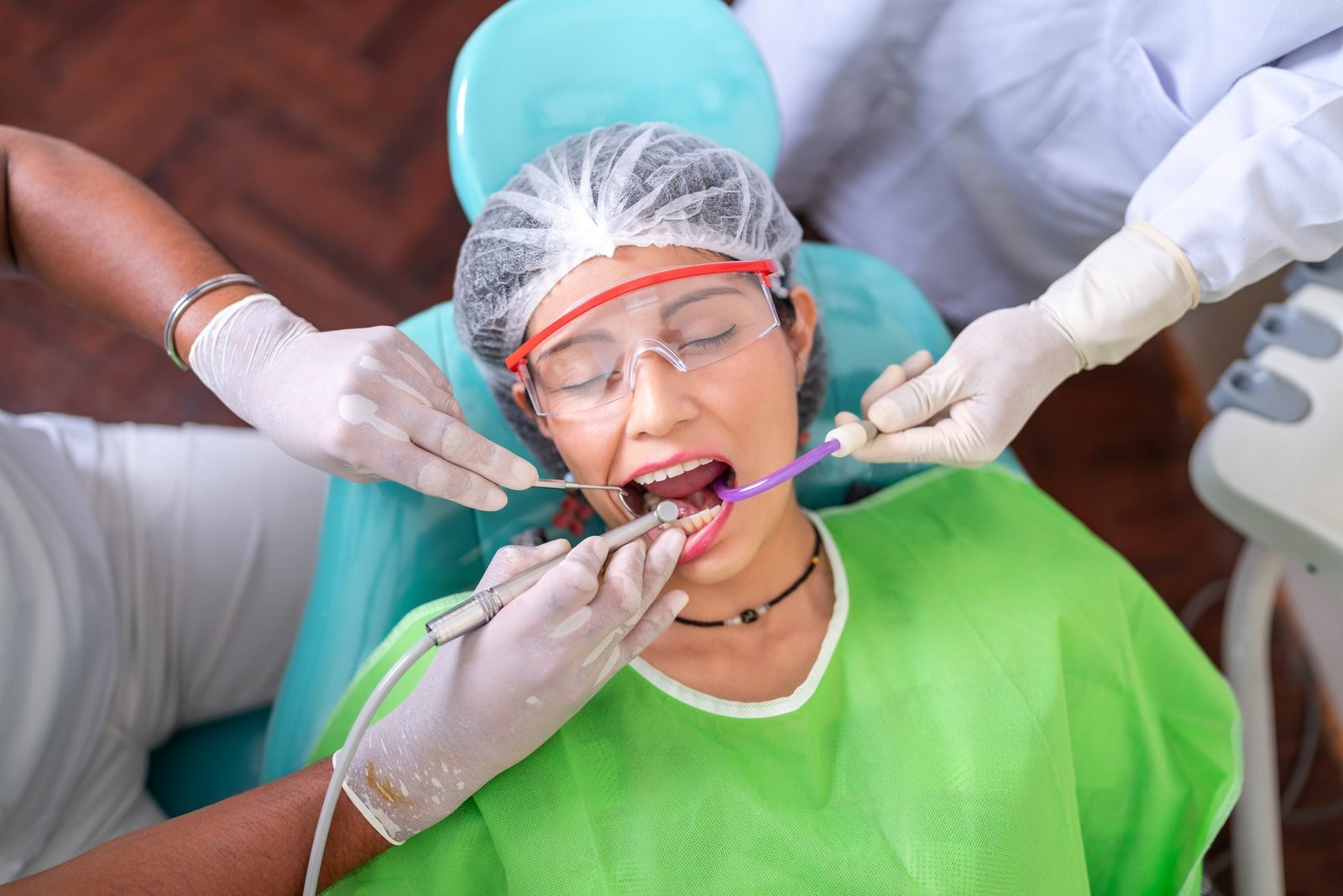 A high-angle close-up photo of a patient in a dental chair wearing safety glasses and a gown, with two gloved medical professionals using dental instruments (a drill and suction/mirror) inside her open mouth during a dental procedure.